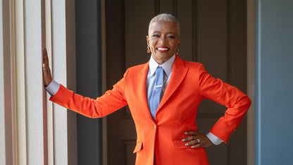 Jennifer wearing a bright orange suit standing in a room with a window and door.